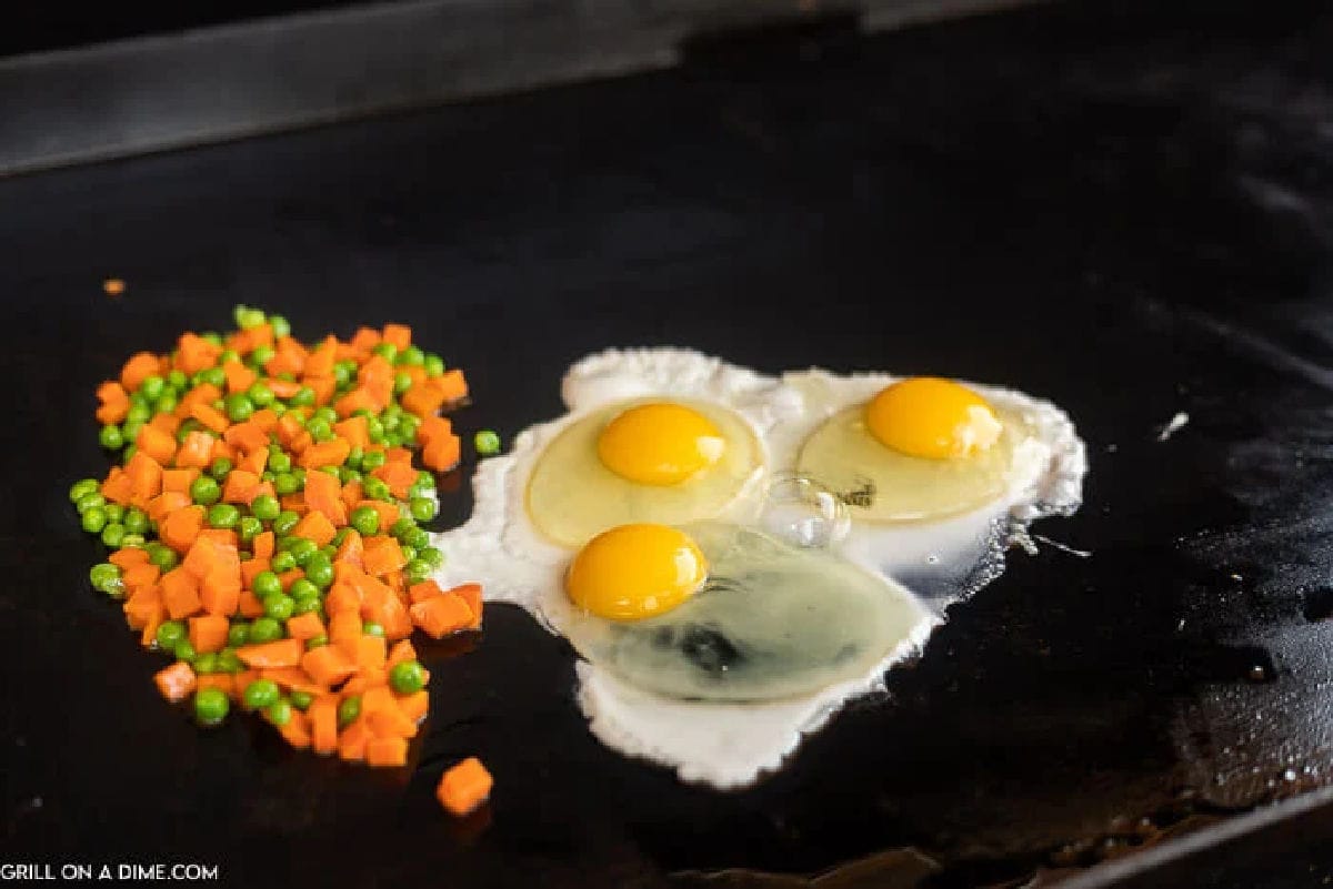 Three raw eggs and a mix of diced carrots and peas are sizzling on a flat-top griddle.