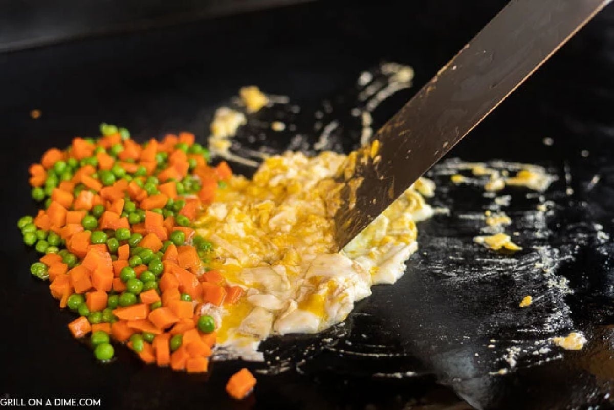 A metal spatula stirs scrambled eggs on a flat griddle beside cooked diced carrots and green peas.