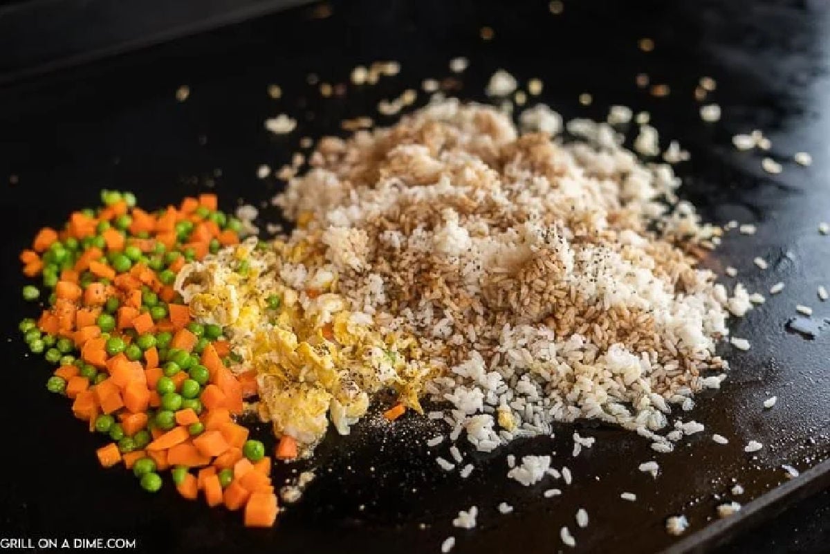 Chopped carrots, peas, scrambled eggs, and cooked rice are being stir-fried together on a flat-top griddle.