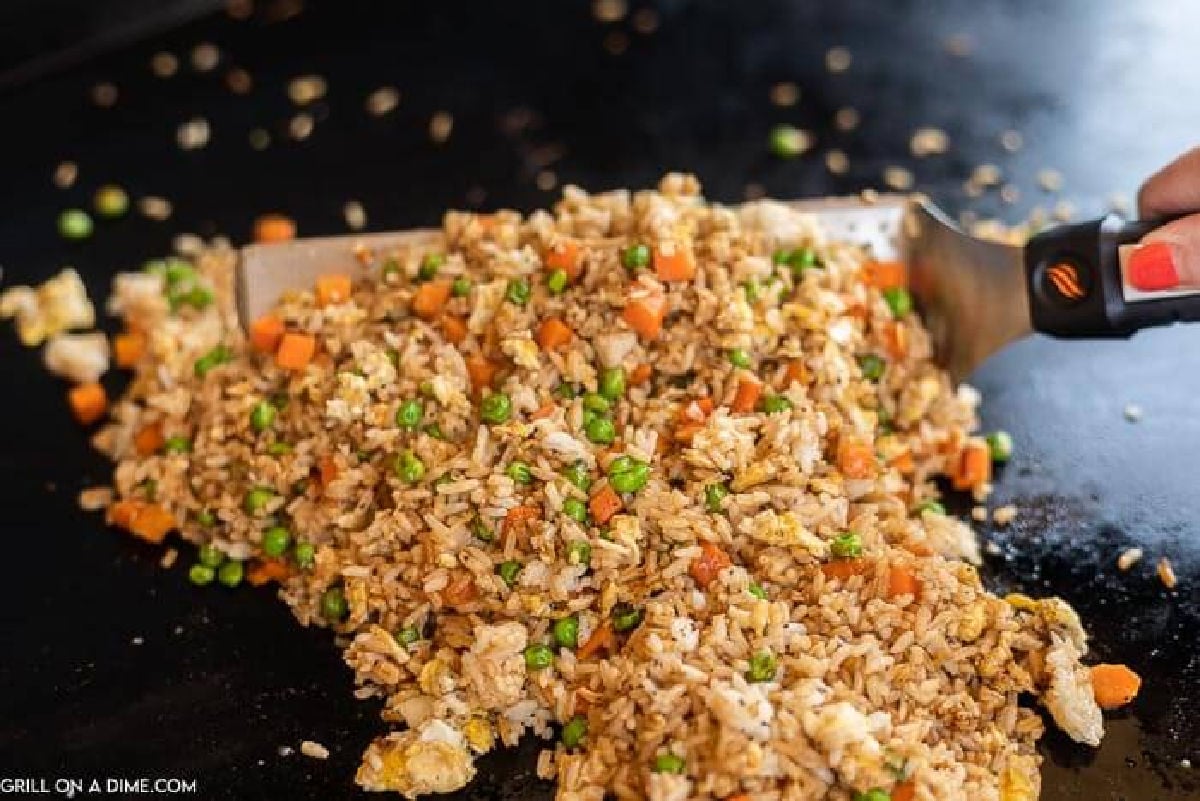 A close-up of freshly cooked Fried Rice with peas, carrots, and scrambled eggs being scooped with a spatula on a flat grill.