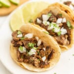 Two Blackstone Street Tacos filled with grilled beef, diced onions, and chopped cilantro sit on a plate with lime slices in the background.