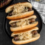 Grilled Bratwurst topped with sauerkraut and placed in hot dog buns on a black plate.
