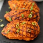 Three Grilled Buffalo Chicken breasts with visible char marks, coated in a glossy sauce, and garnished with chopped green onions, arranged on a dark slate serving board.