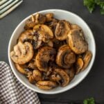 A white bowl filled with Blackstone Sautéed Mushrooms, seasoned with herbs, sits on a dark surface next to a checkered cloth and a fork, with fresh parsley in the background.