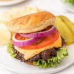 Enjoy our Blackstone Cheeseburgers topped with lettuce, tomato, red onion, pickles, and melted cheese on a soft bun. Served on a white plate with crispy potato chips and extra pickle slices on the side.