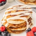 Cinnamon Roll Pancakes stacked on a plate topped with a glaze with a side of fresh berries