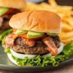 A Blackstone Avocado Bacon Burger with lettuce, onion, tomato, and avocado slices in a soft bun, served on a plate with French fries in the background.