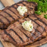 Two steaks with butter and parsley on a cutting board.