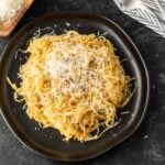 A black plate filled with Spaghetti Squash cooked on a Blackstone Griddle is topped with freshly grated Parmesan cheese, set on a dark textured surface with a fork and napkin nearby.