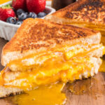 Grilled Cheese that is cut in half and the melted cheese is spread over the wooden board with fresh fruit in the background