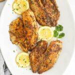 Four seasoned and cooked Blackstone Blackened Tilapia fillets garnished with chopped herbs, served with lemon slices and cilantro on a white plate.