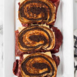 Three Blackstone Reuben Sandwiches made with swirled rye bread are stacked vertically on a white rectangular plate, showcasing layers of corned beef, cheese, and other traditional fillings.