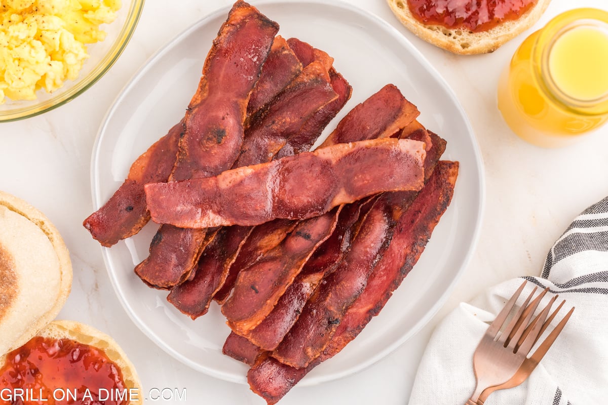 Cooked Turkey Bacon stacked on a plate with scrambled eggs and English Muffins on the side with a side of orange juice