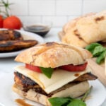 A chicken caprese sandwich with cheese, tomato, and basil on ciabatta bread sits on a white plate, with fresh tomatoes, basil, and another sandwich in the background.