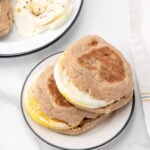 Two breakfast sandwiches with fried eggs and egg patties on whole wheat English muffins are stacked on a plate. A plate with a cooked egg is partially visible in the background.
