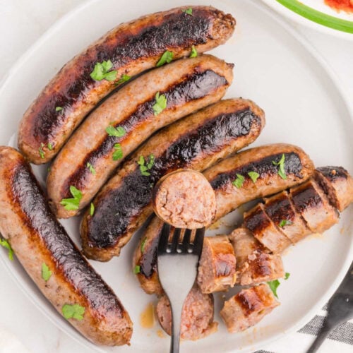 A white plate with grilled Italian sausage on Blackstone, garnished with parsley. One sausage is sliced, and a fork holds a piece above the plate. The sausages have visible grill marks.