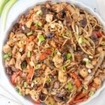 A plate of Chicken Lo Mein on Blackstone features noodles, juicy chicken pieces, mushrooms, red bell peppers, onions, and green onions, garnished with sesame seeds. Chopsticks and fresh green onions sit beside the plate.
