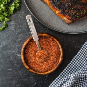 A bowl of dry spice rub with a metal tablespoon sits on a dark surface next to coffee rubbed steak, fresh parsley, and a blue and white checkered cloth.