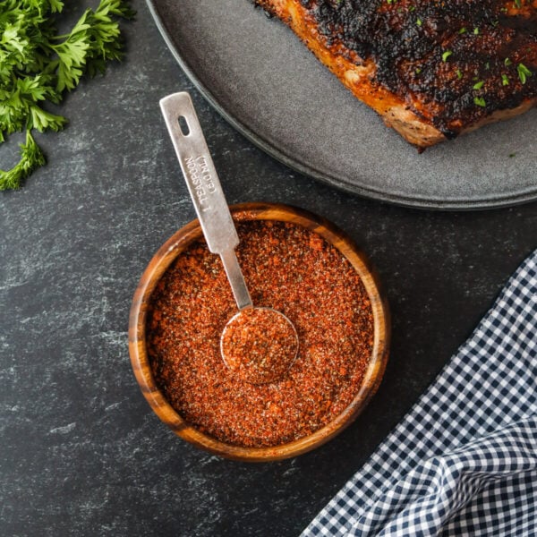 A bowl of dry spice rub with a metal tablespoon sits on a dark surface next to coffee rubbed steak, fresh parsley, and a blue and white checkered cloth.