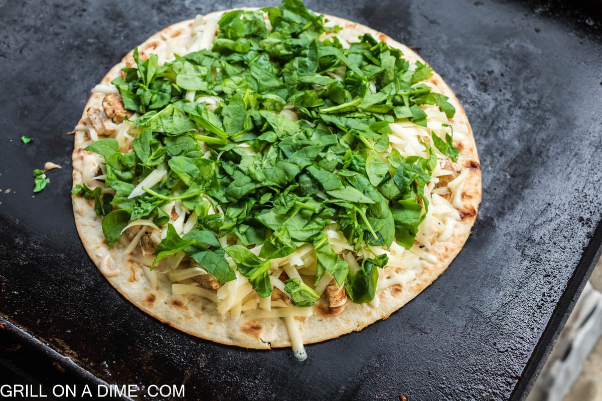 pizza topped with shredded spinach shredded cheese and chicken on the blackstone griddle