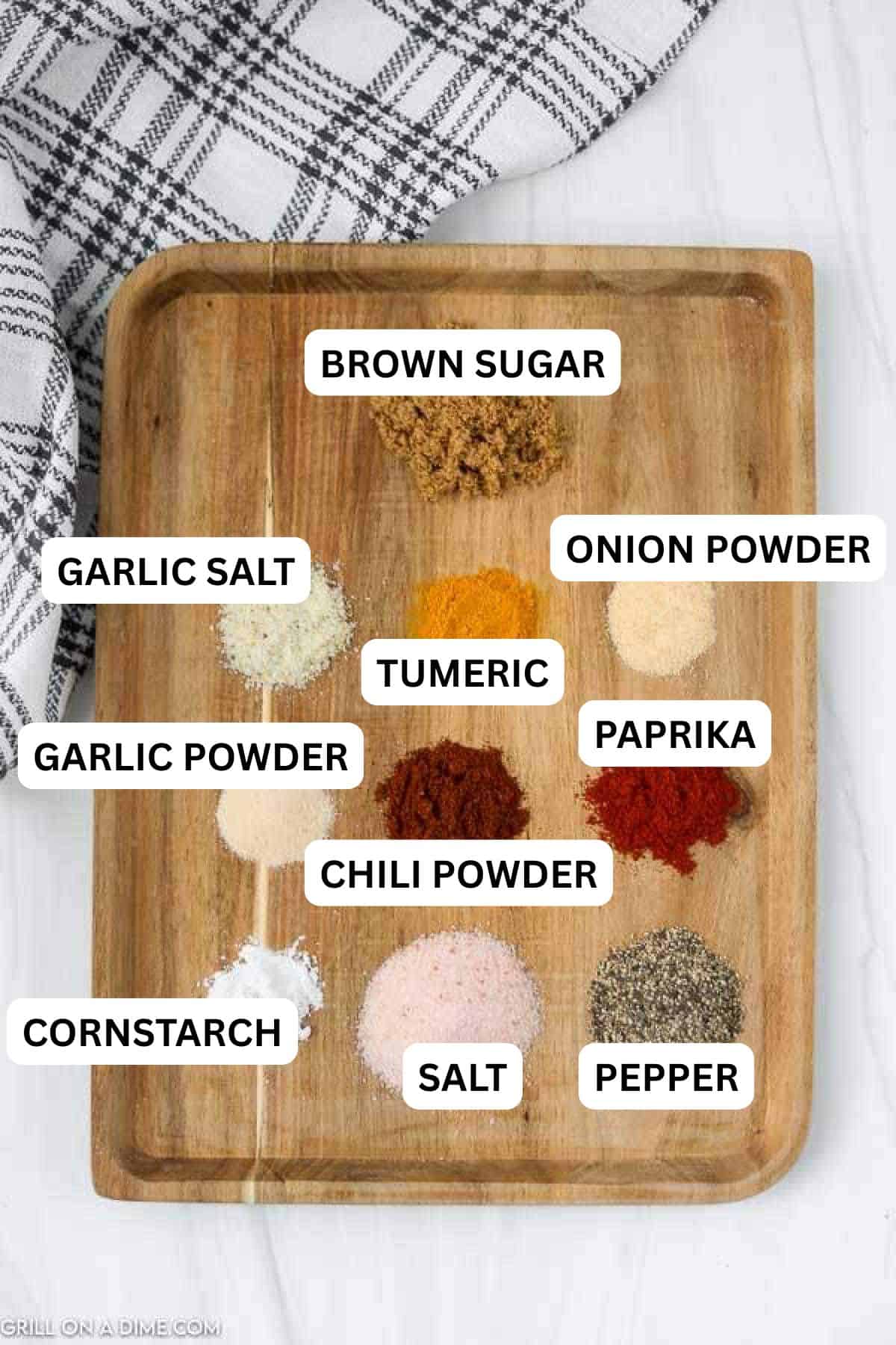 A wooden board displays labeled piles of Texas Roadhouse Steak Seasoning, brown sugar, garlic salt, cornstarch, turmeric, chili powder, salt, onion powder, paprika, and pepper. A checkered towel adds a cozy touch in the corner.