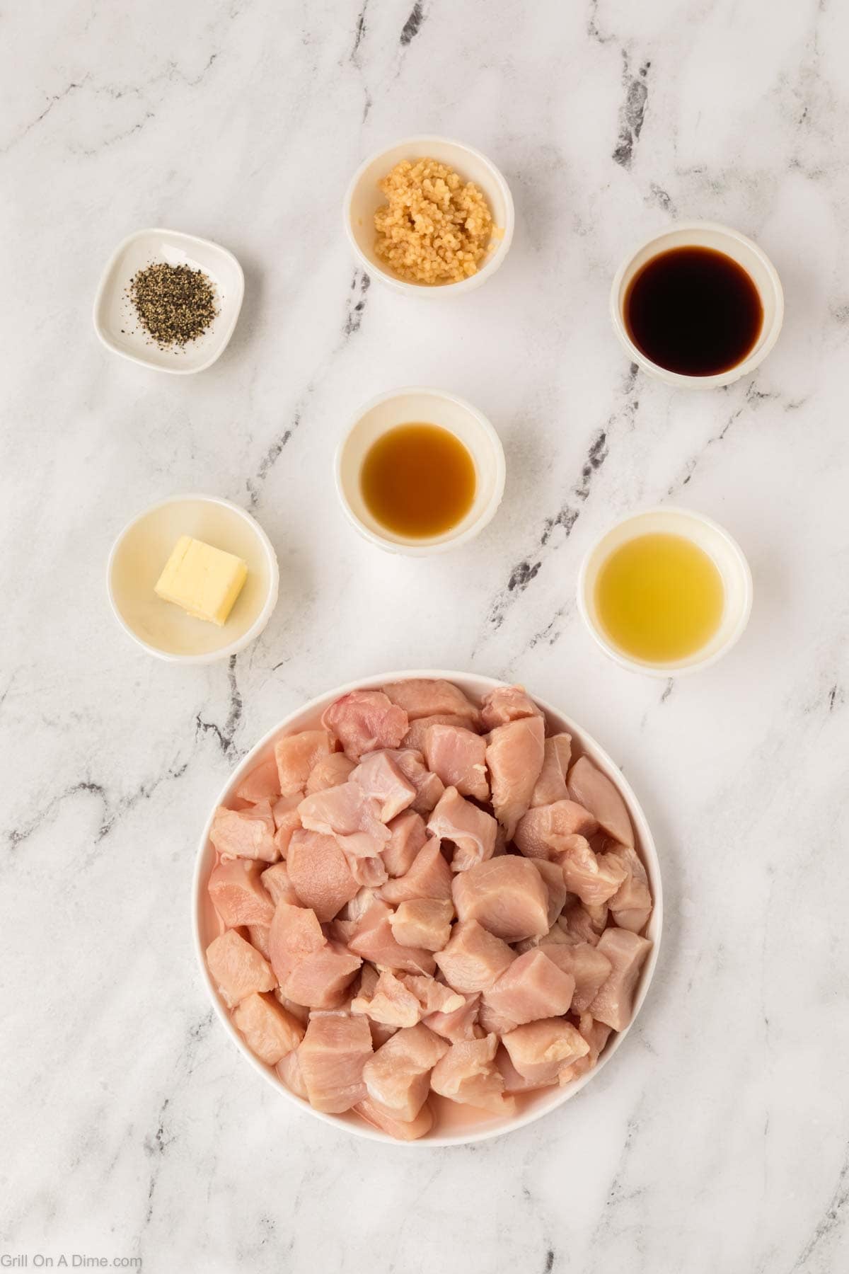A plate of raw cubed chicken for Hibachi Chicken is surrounded by small bowls of minced garlic, black pepper, soy sauce, butter, honey, and apple cider vinegar, all neatly arranged on a white marble surface.