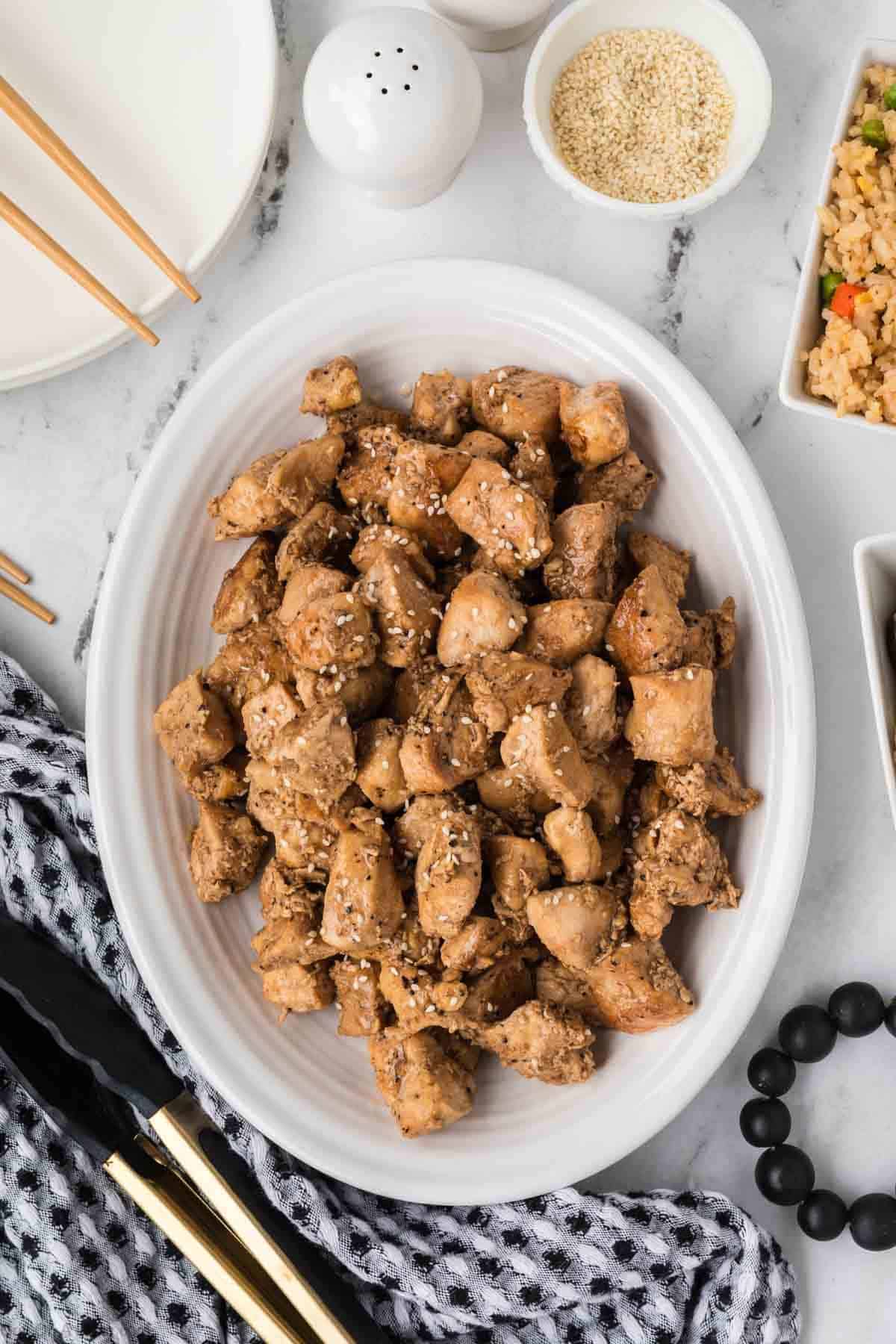A white oval plate filled with bite-sized pieces of Hibachi Chicken sprinkled with sesame seeds, surrounded by chopsticks, a patterned towel, tongs, and small bowls of rice and seasonings on a marble surface.