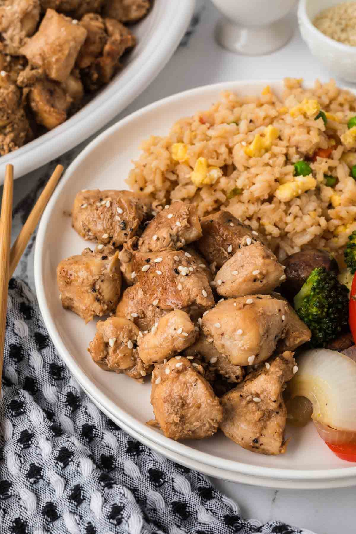 A plate of Hibachi Chicken grilled chunks topped with sesame seeds, served with fried rice, broccoli, red bell pepper, and onions. Chopsticks rest on the side, and a patterned napkin is underneath the plate.