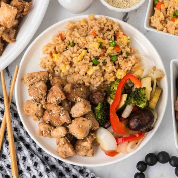A plate of Hibachi Chicken with sesame seeds, fried rice with peas and carrots, and sautéed vegetables—broccoli, mushrooms, red bell peppers, and onions. Chopsticks and side dishes are nearby.