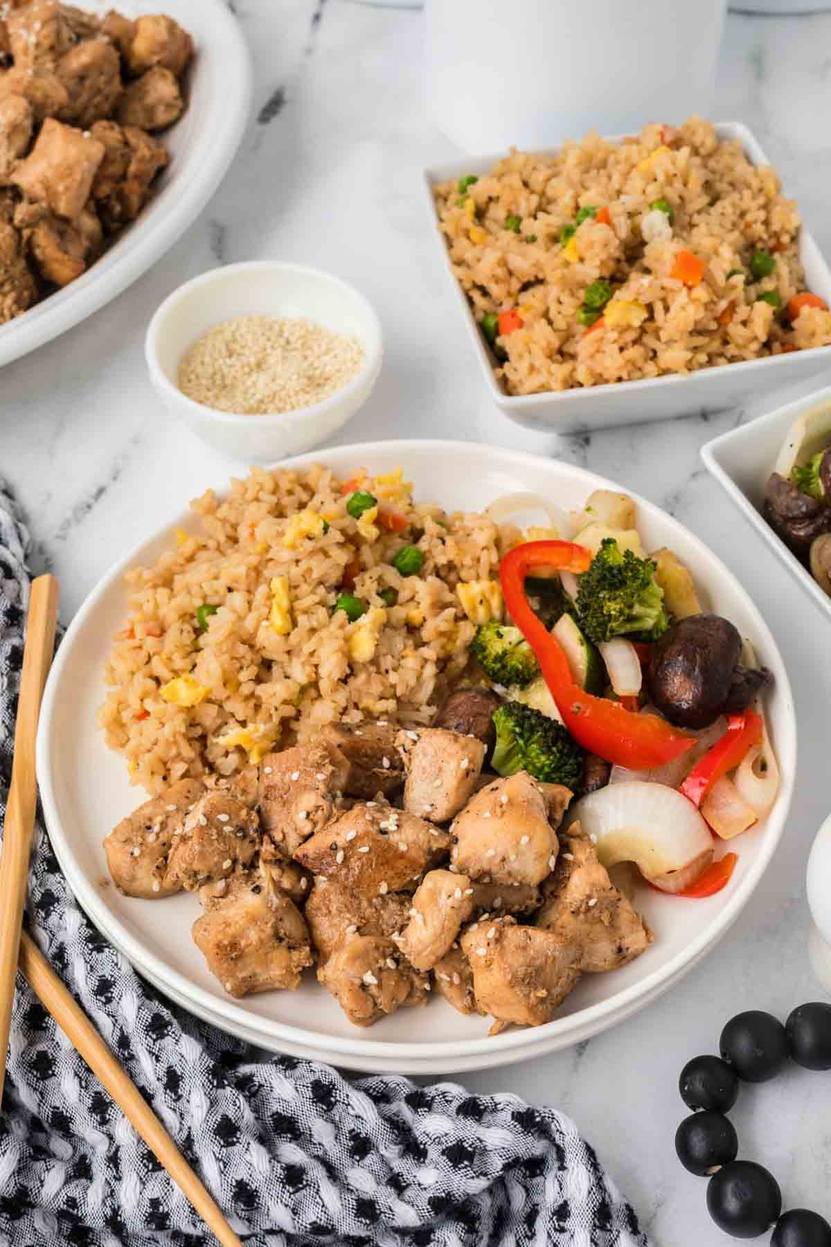 A plate with Hibachi Chicken—grilled chicken pieces topped with sesame seeds—served alongside fried rice and stir-fried vegetables like broccoli, red bell pepper, onions, and mushrooms. Fried rice and sesame seeds are in bowls nearby. Chopsticks rest on the side.