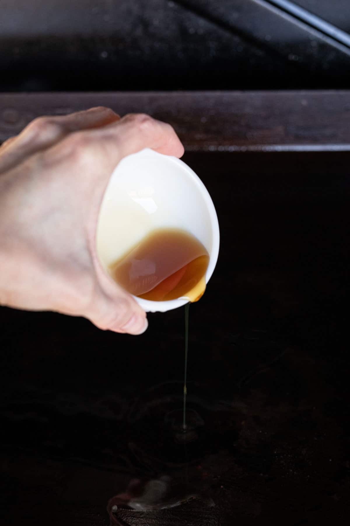 A hand pours a small amount of brown liquid from a white cup onto a dark surface, likely for cooking Hibachi Chicken.