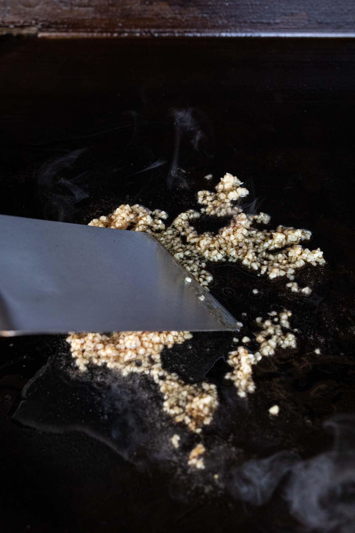 A metal spatula is scraping minced garlic on a hot, dark griddle alongside sizzling Hibachi Chicken, with steam rising from the cooking surface.