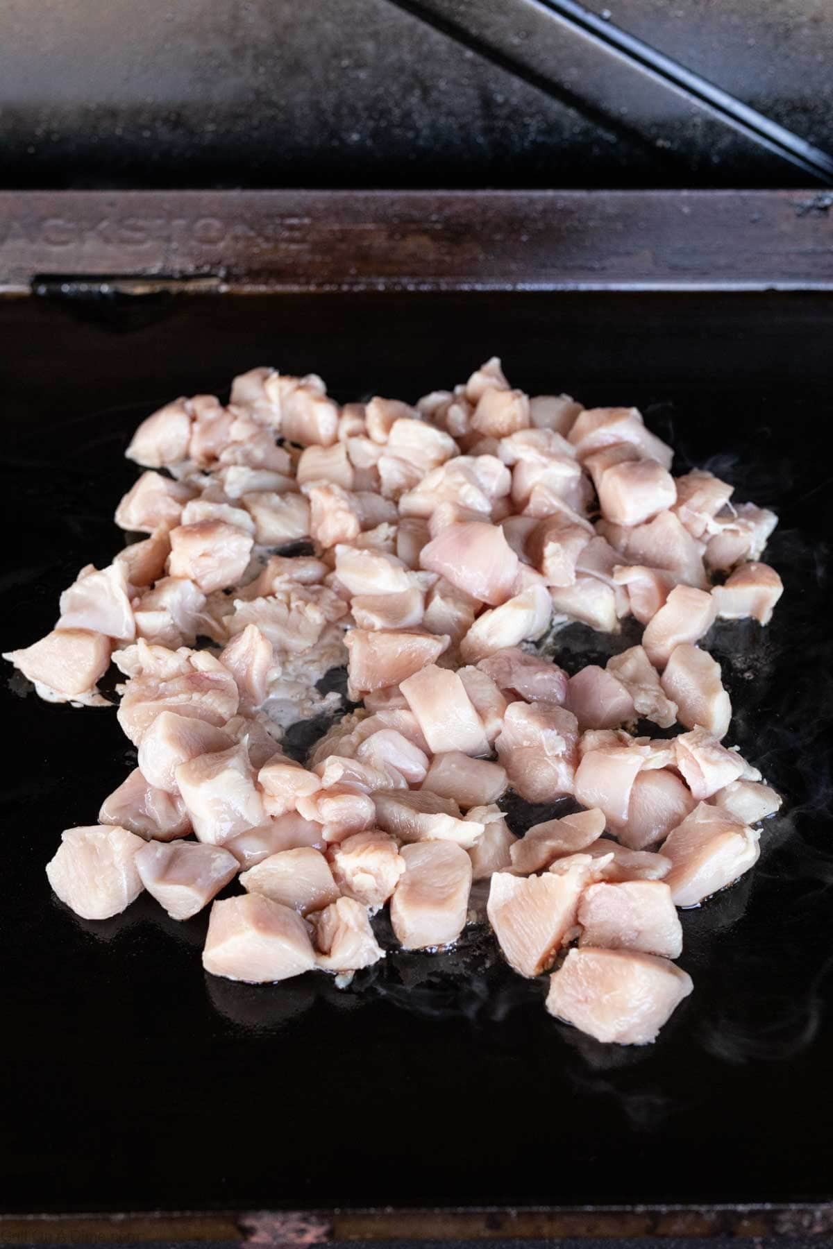 Hibachi Chicken: Diced raw chicken pieces cooking on a flat-top griddle, beginning to turn white as they heat up.