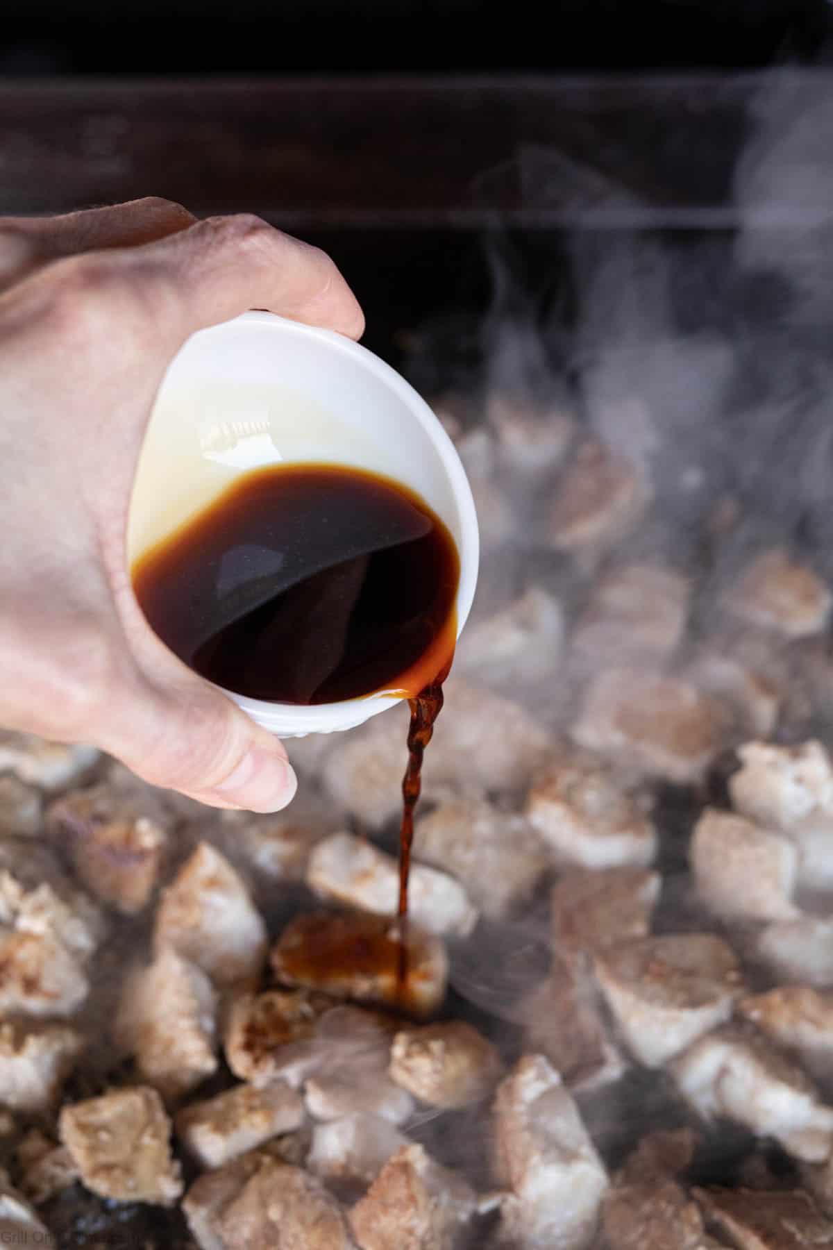 A hand pours a dark sauce from a white cup onto sizzling, cubed Hibachi Chicken cooking on a hot, steamy surface.