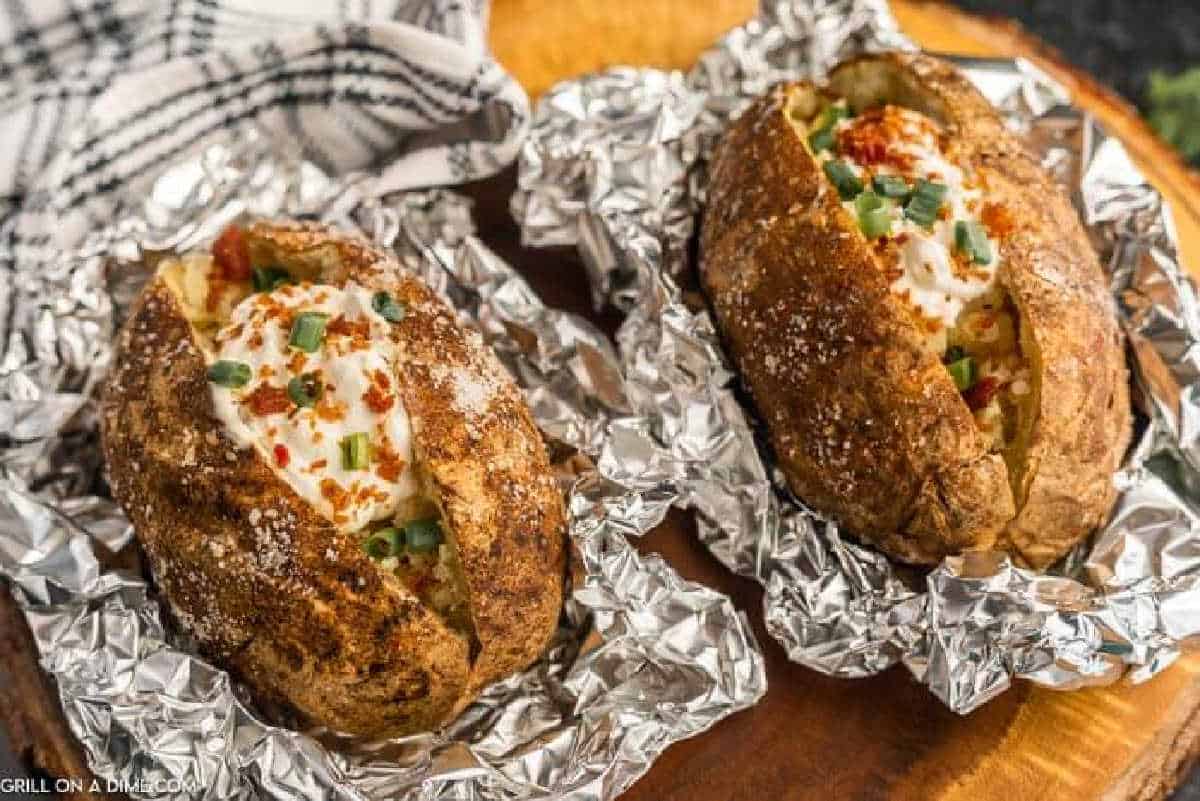 Two baked potatoes wrapped in foil, are topped with sour cream, chopped green onions, crumbled bacon, and paprika on a wooden surface with a checkered cloth nearby.