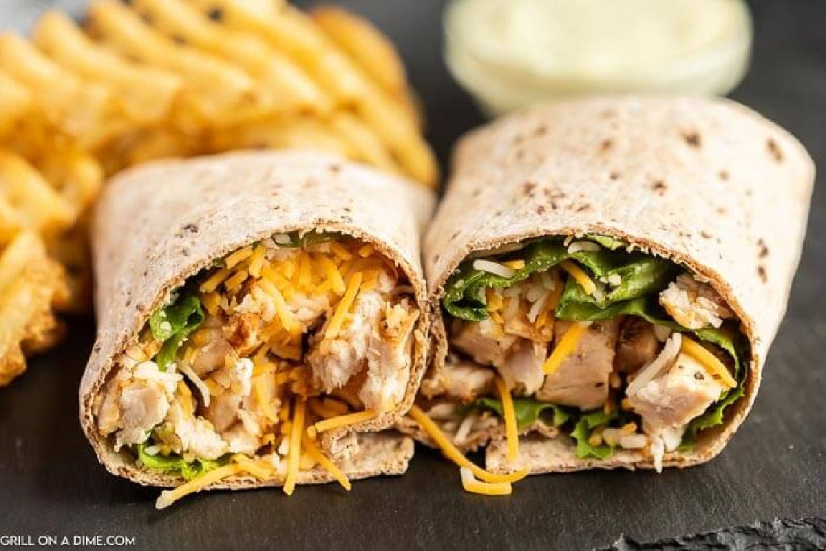 A close-up of a Chick-Fil-A Cool Wrap filled with grilled chicken, shredded cheddar cheese, and leafy greens, served alongside crispy waffle fries and a dipping sauce in the background.