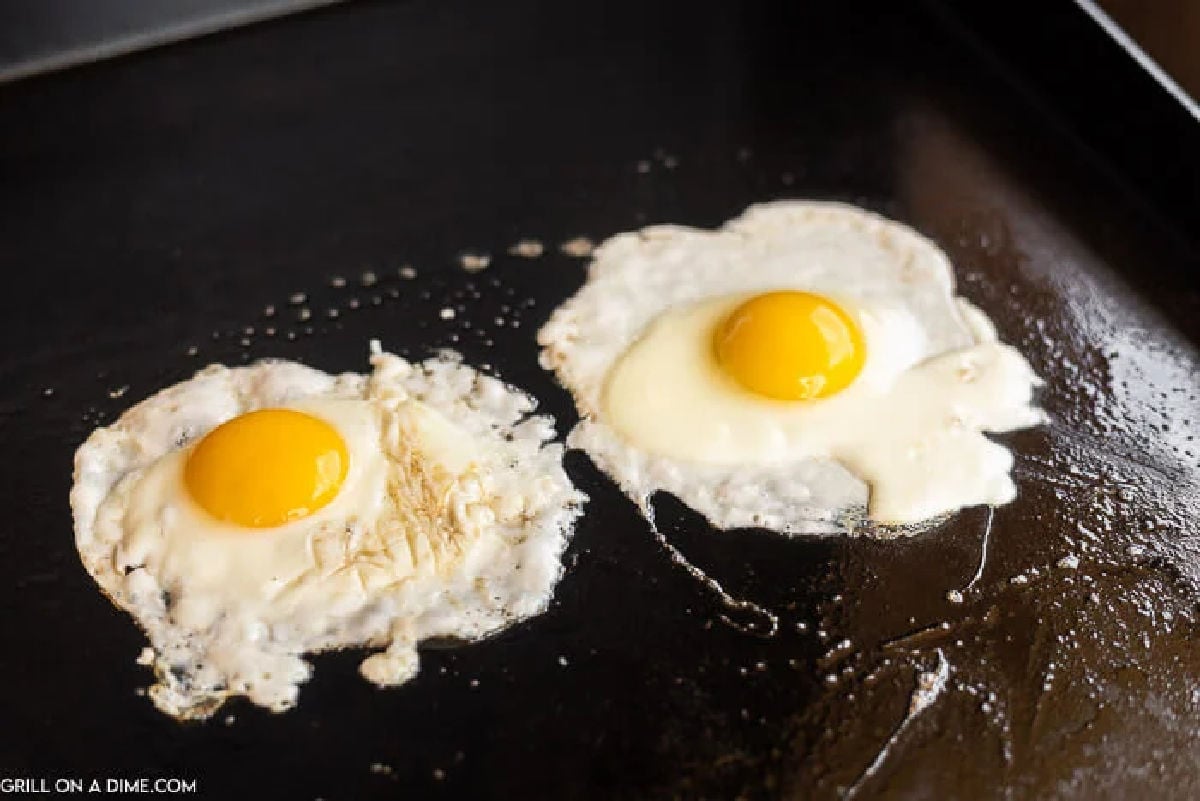 Two sunny-side-up fried eggs on a Blackstone griddle, with bright yolks intact and whites gently spreading out.