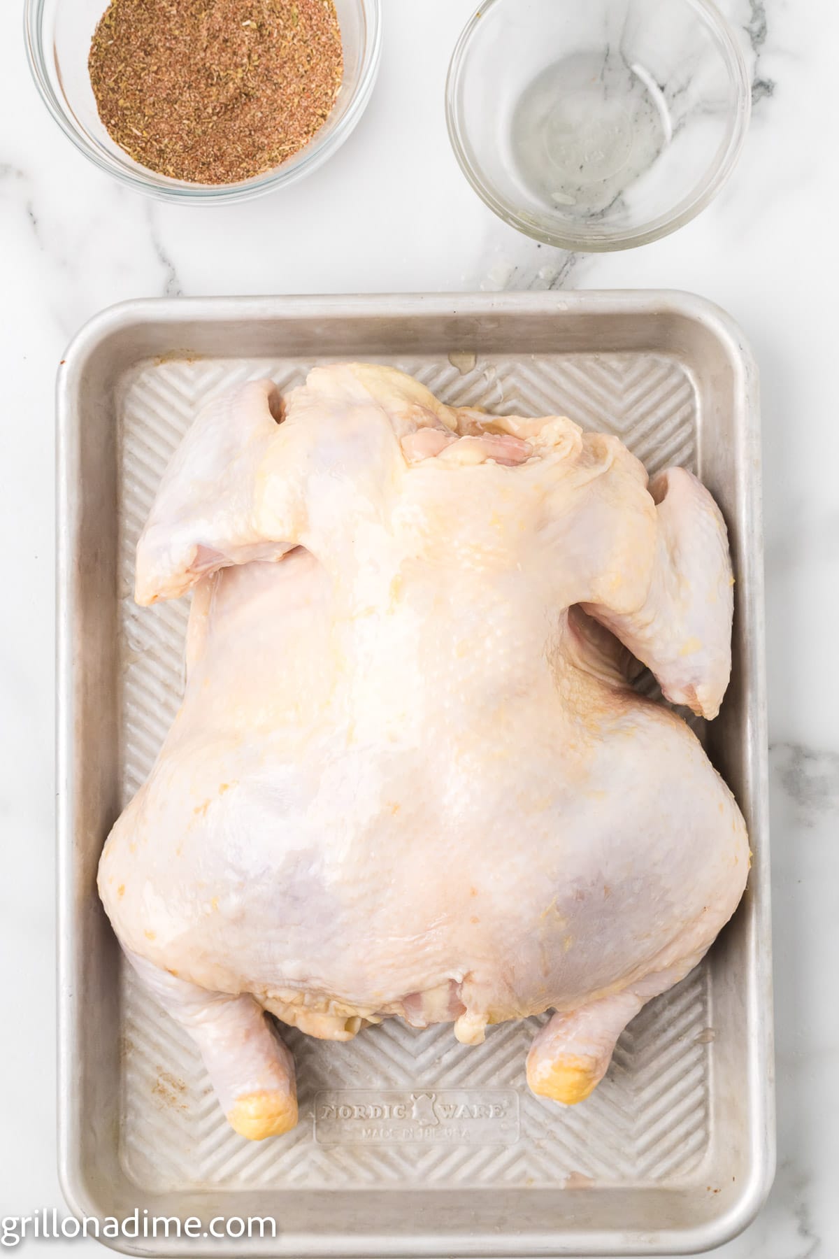 Whole chicken on a baking sheet with a small bowls of seasoning and oil
