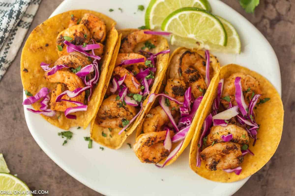 4 Shrimp Tacos in corn tortillas topped with red cabbage and chopped cilantro on a plate with a side of lime wedges.