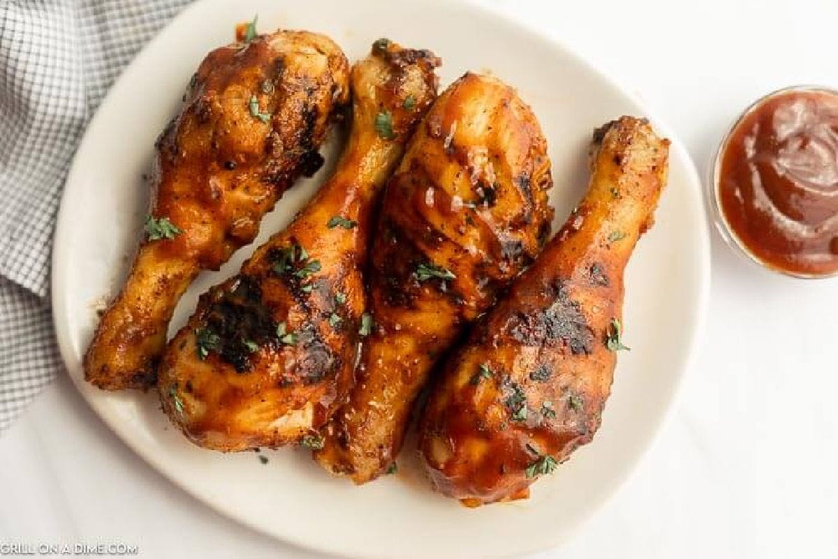 Four Grilled BBQ Chicken Drumsticks garnished with herbs are arranged on a white plate, accompanied by a small bowl of barbecue sauce and a checkered napkin nearby.