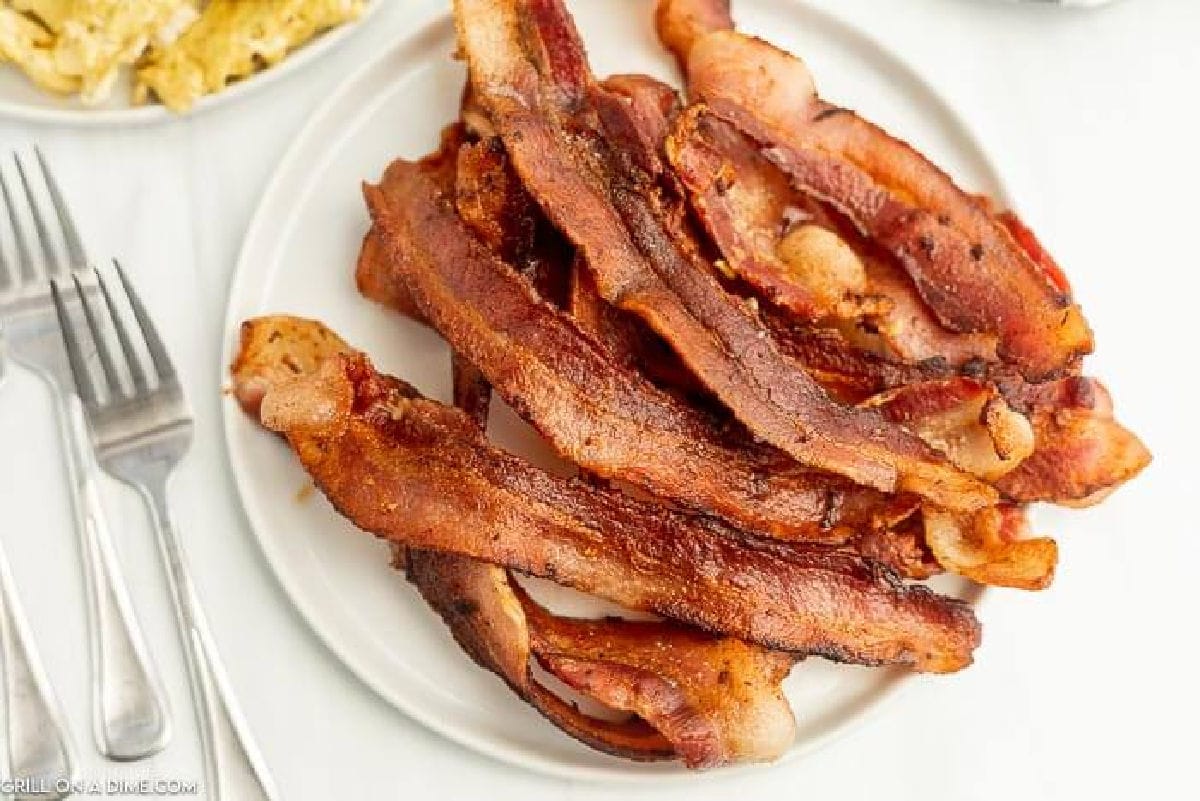 A white plate piled with crispy, cooked bacon strips sits on a white surface next to forks and a dish of scrambled eggs.