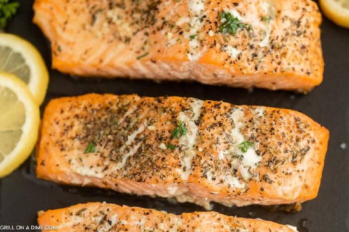 Three Olive Garden Herb Grilled Salmon fillets topped with herbs and melted butter are arranged on a dark surface, with lemon slices placed nearby.