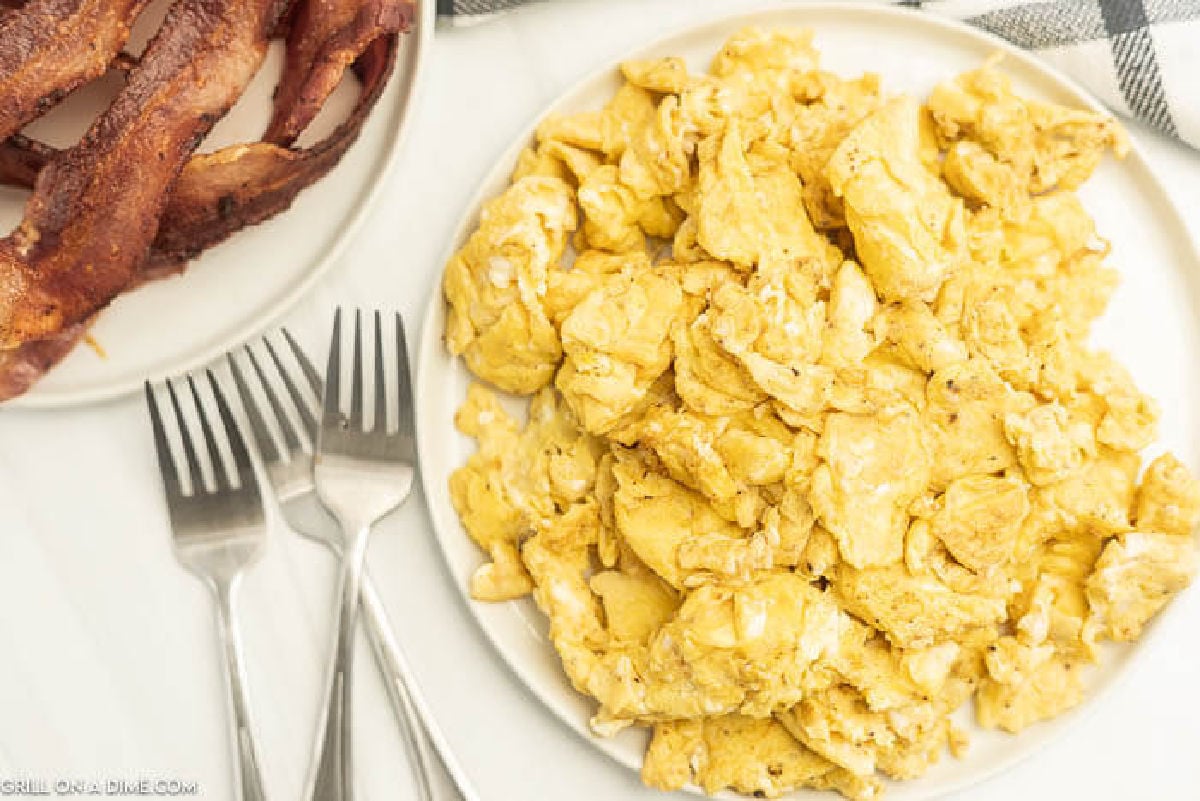 A plate of Blackstone Scrambled Eggs sits next to cooked bacon, with three forks beside the plates on a white surface. A checkered cloth is partly visible in the background.