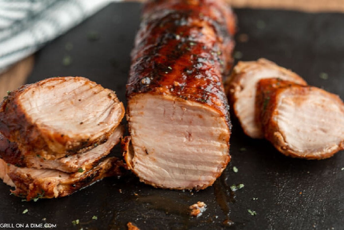 Sliced grilled bacon wrapped pork tenderloin on a dark cutting board, showing a juicy, well-cooked interior with a seasoned, browned exterior. Some slices are arranged around the main piece.