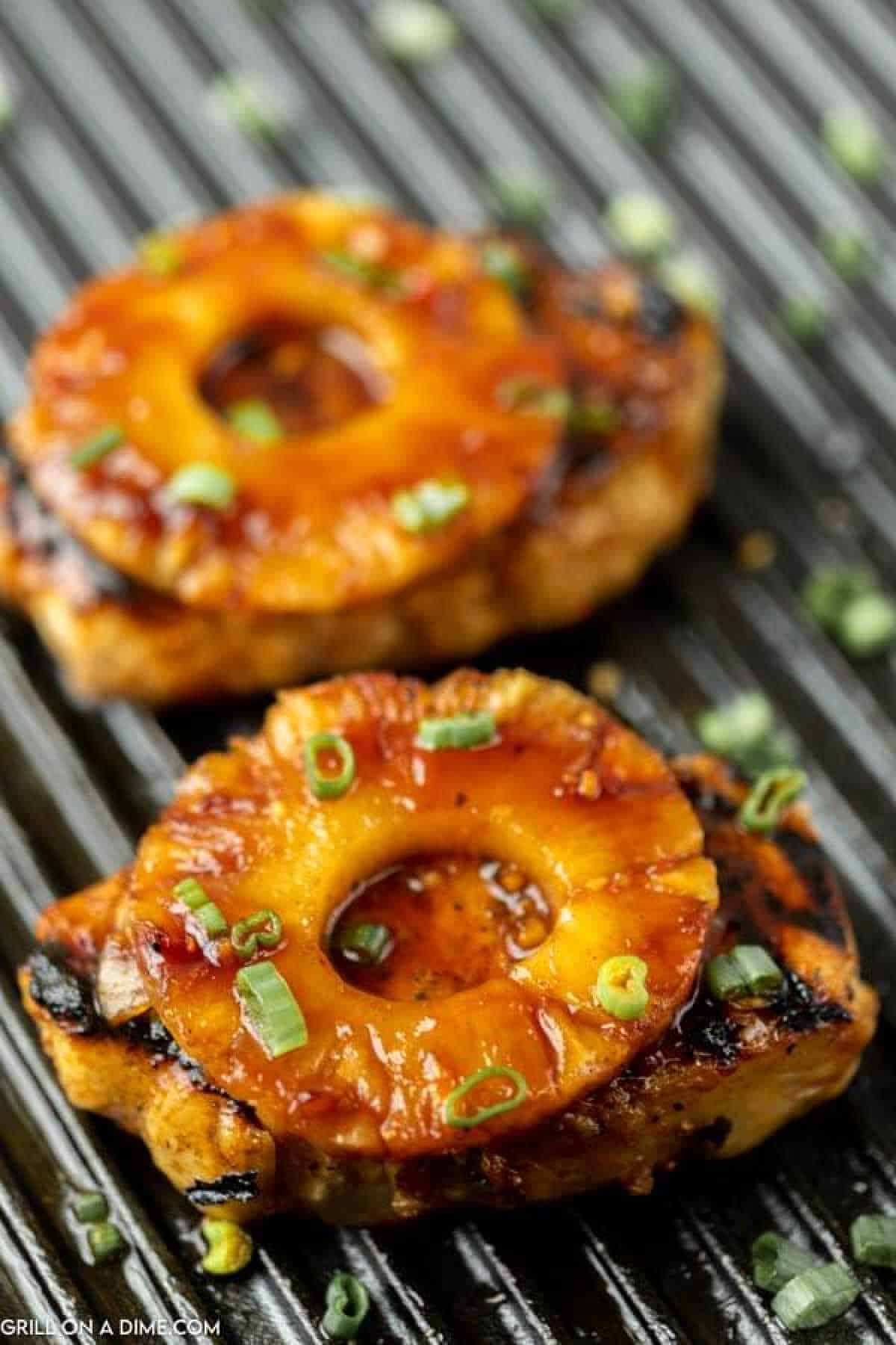 Two grilled pork chops topped with caramelized pineapple rings and chopped green onions, sitting on a ridged grill pan.