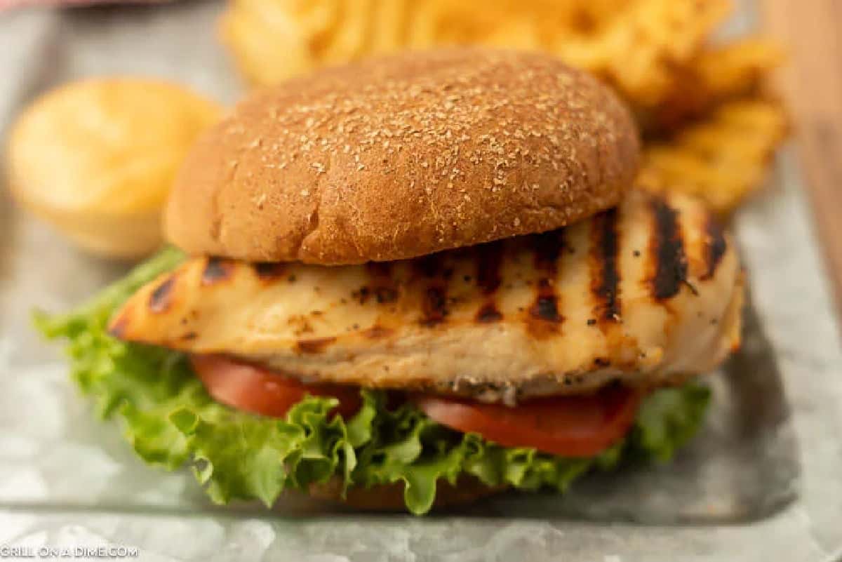 A Chick-Fil-A Grilled Chicken Sandwich with lettuce and tomato on a whole wheat bun, served alongside crispy waffle fries and a biscuit on a metal tray.