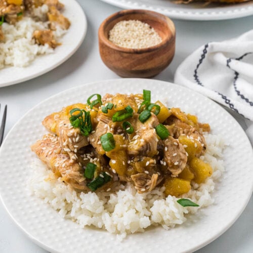 A plate of white rice topped with Blackstone pineapple chicken, sliced green onions, and sprinkled with sesame seeds. A small bowl of sesame seeds and another plate of food are in the background.
