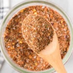 A wooden spoon holding a heap of mixed dry spices and herbs, including Montreal steak seasoning, rests over a glass jar filled with the same flavorful blend.
