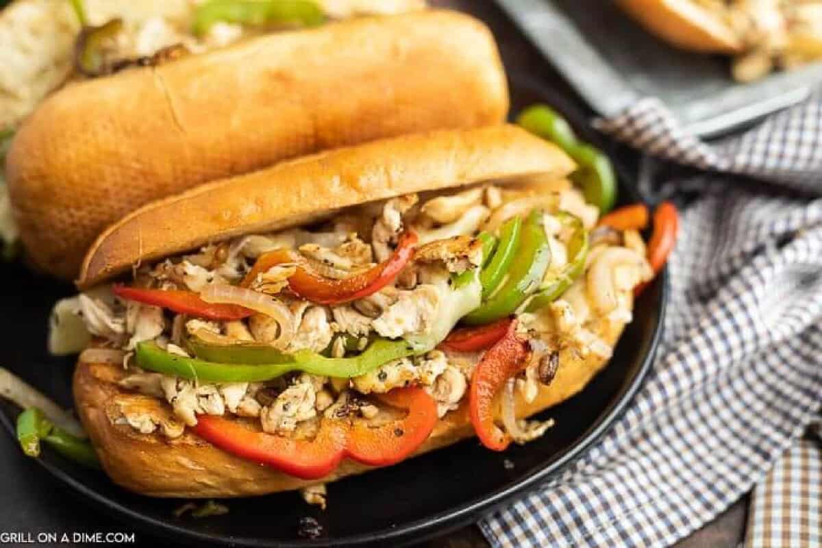 A close-up of a Blackstone Chicken Philly Cheesesteak, filled with shredded grilled chicken, sautéed green and red bell peppers, and onions on a toasted hoagie roll, served on a black plate with a checked napkin nearby.