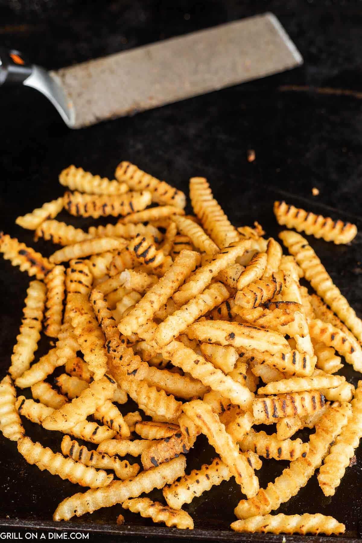 A pile of grilled crinkle-cut frozen French fries on a dark Blackstone grill surface, with a metal spatula in the background. 